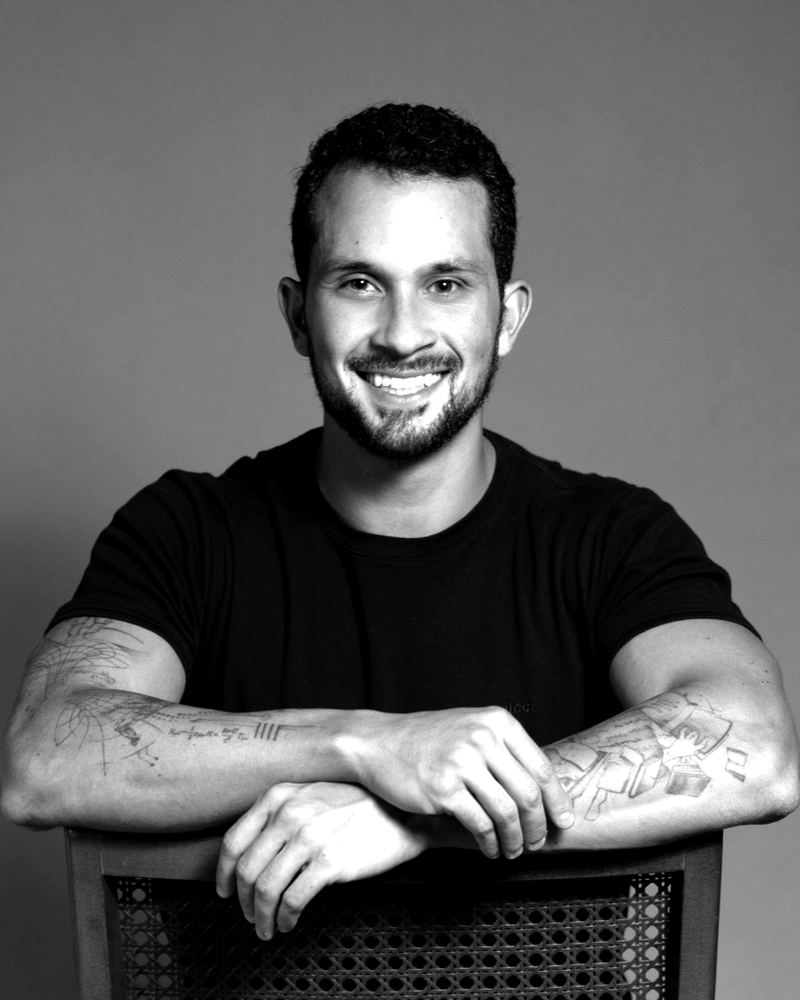 Matheus Gomes, em preto e branco, sorrindo e apoiado no encosto de uma cadeira